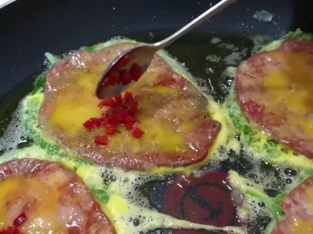 Primer plano de una cuchara añadiendo chiles verdes y rojos picados sobre las tortitas de ternera que se cocinan en la sartén.