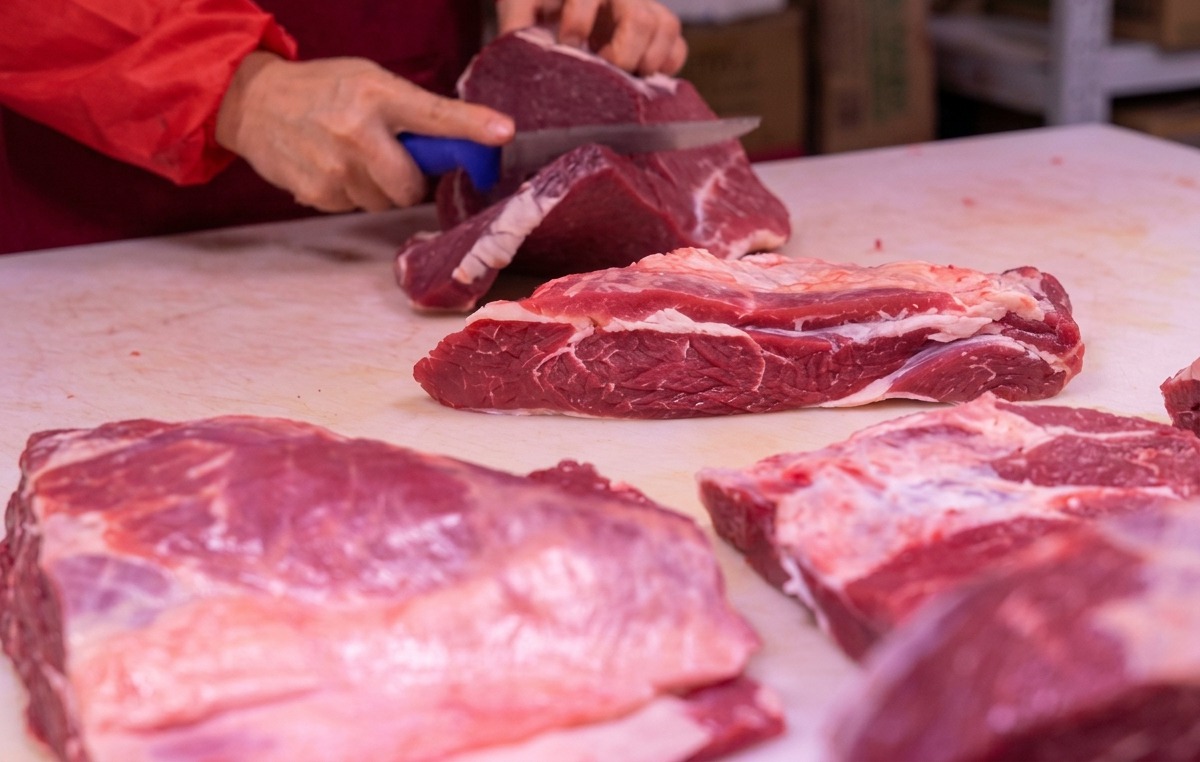Manos usando un cuchillo afilado para cortar carne roja sobre una tabla de cortar blanca.
