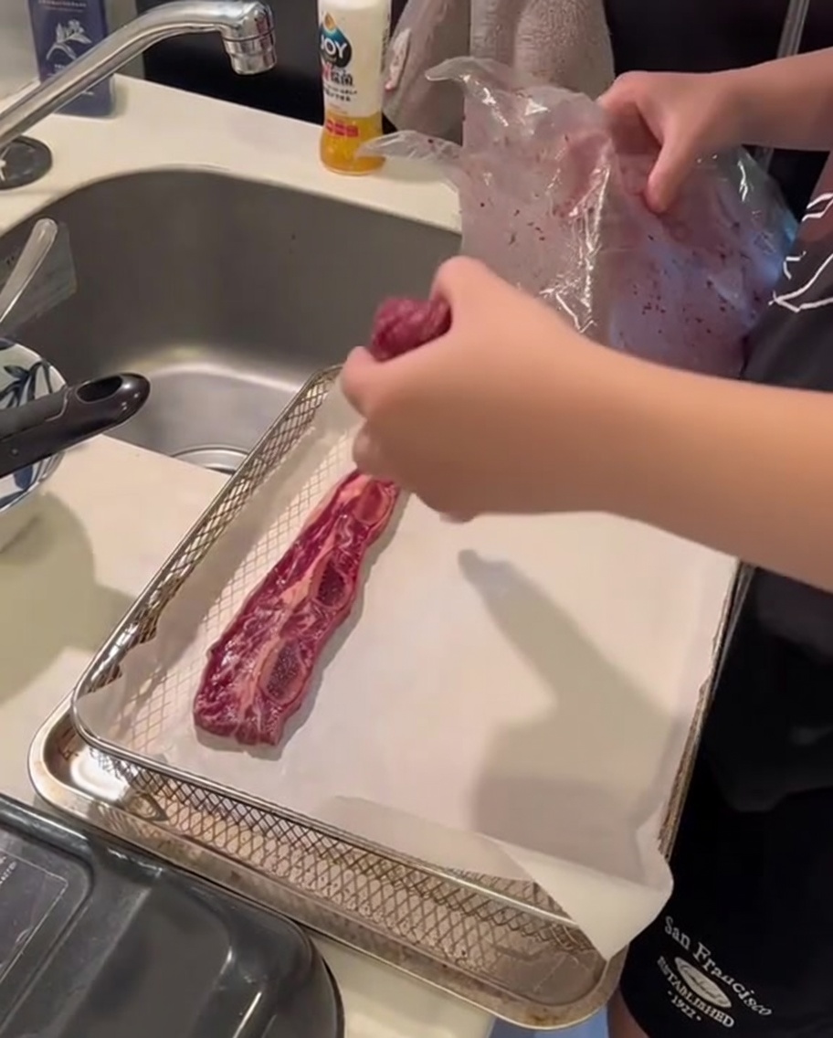 Costillas crudas estilo flanken dispuestas ordenadamente sobre una hoja de papel pergamino dentro de la canasta de una freidora de aire.