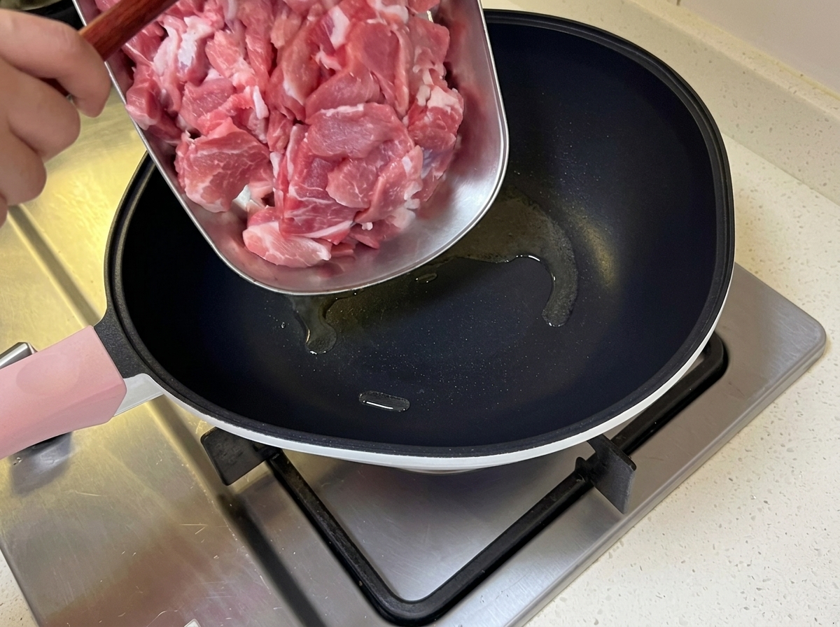 Cerdo cortado en láminas finas siendo añadido a una sartén caliente con aceite brillante.
