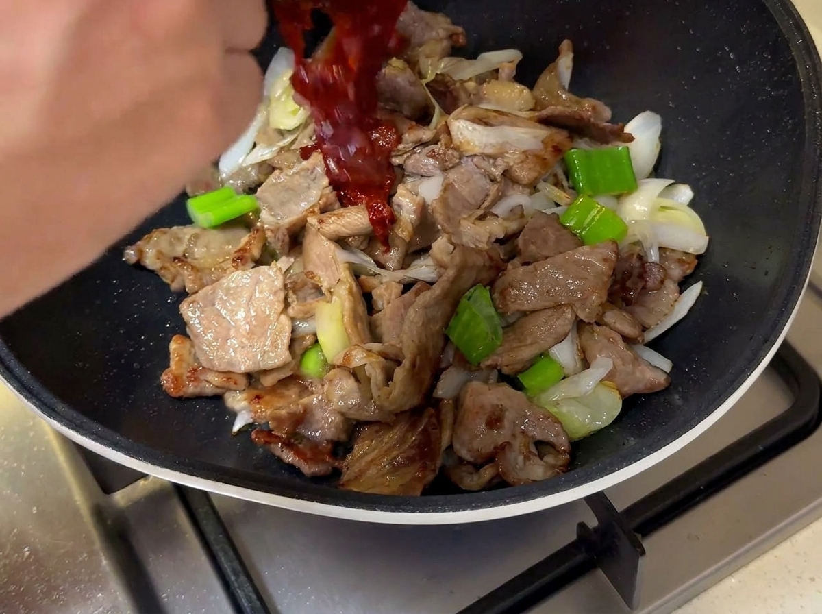 La salsa roja oscura siendo vertida desde un tazón pequeño sobre la carne y las cebolletas en la sartén.