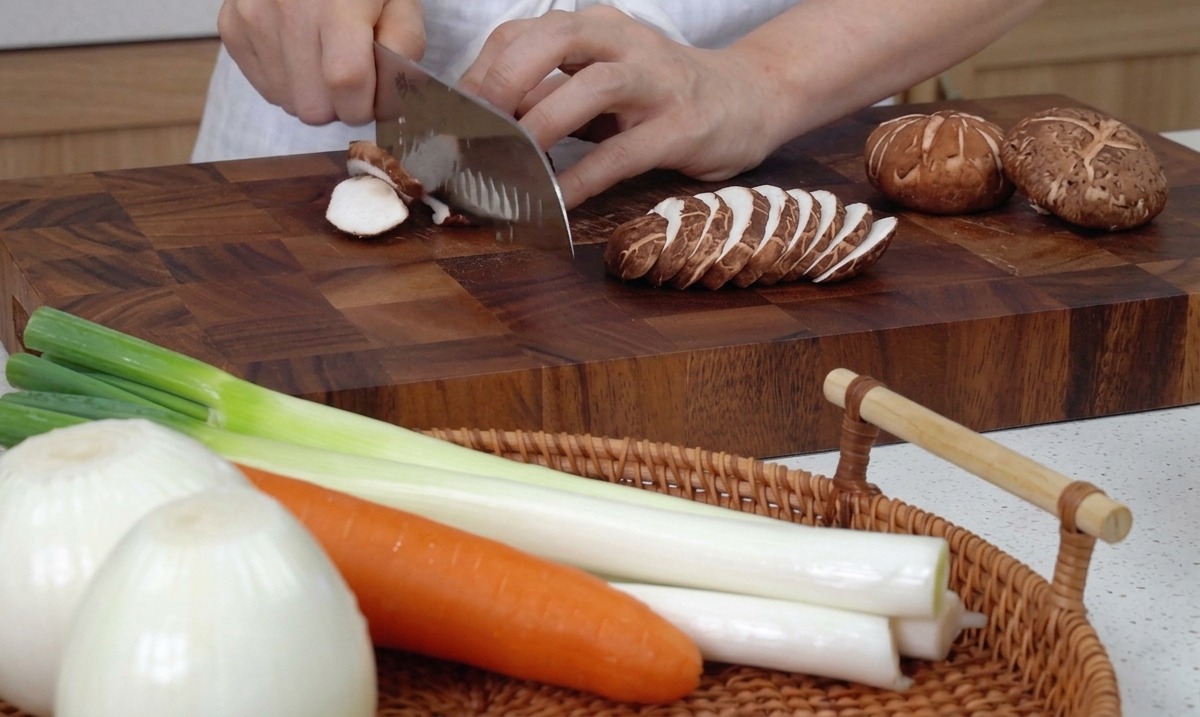 Manos usando un cuchillo grande de cocina para cortar setas shiitake frescas en una tabla de cortar de madera.
