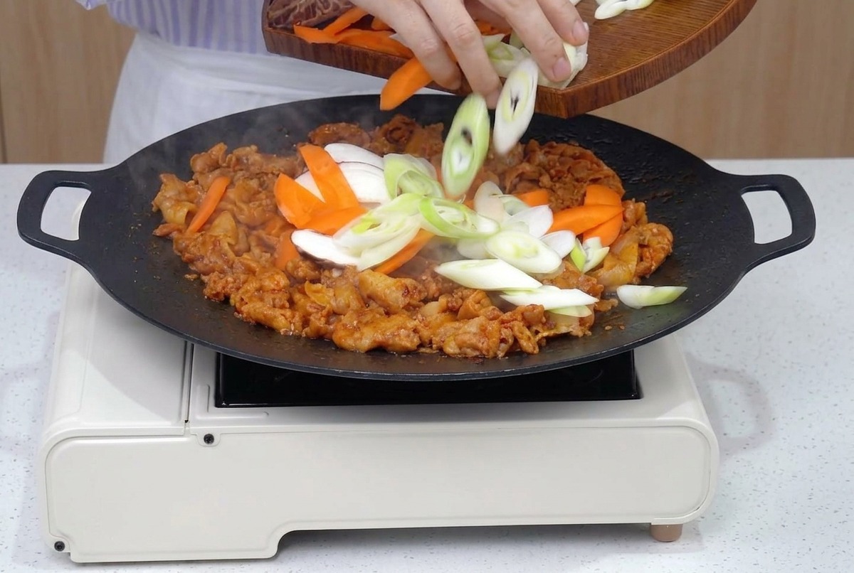 Una sartén llena de panceta de cerdo dorada a la que se unen láminas frescas de zanahorias, cebollas, setas y cebolletas.