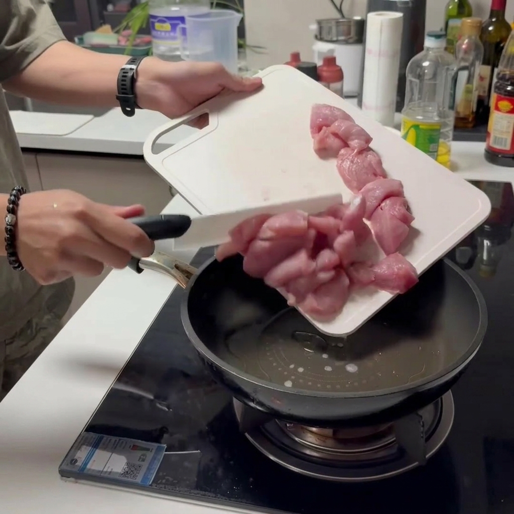Deslizando trozos de cerdo crudo cortado desde una tabla de cortar blanca directamente a una sartén con aceite.