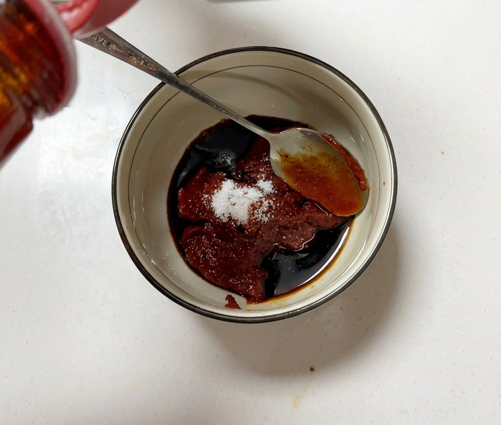 Un tazón pequeño lleno de pasta de chile coreana, azúcar y salsa de soja siendo mezclados con una cuchara de metal.