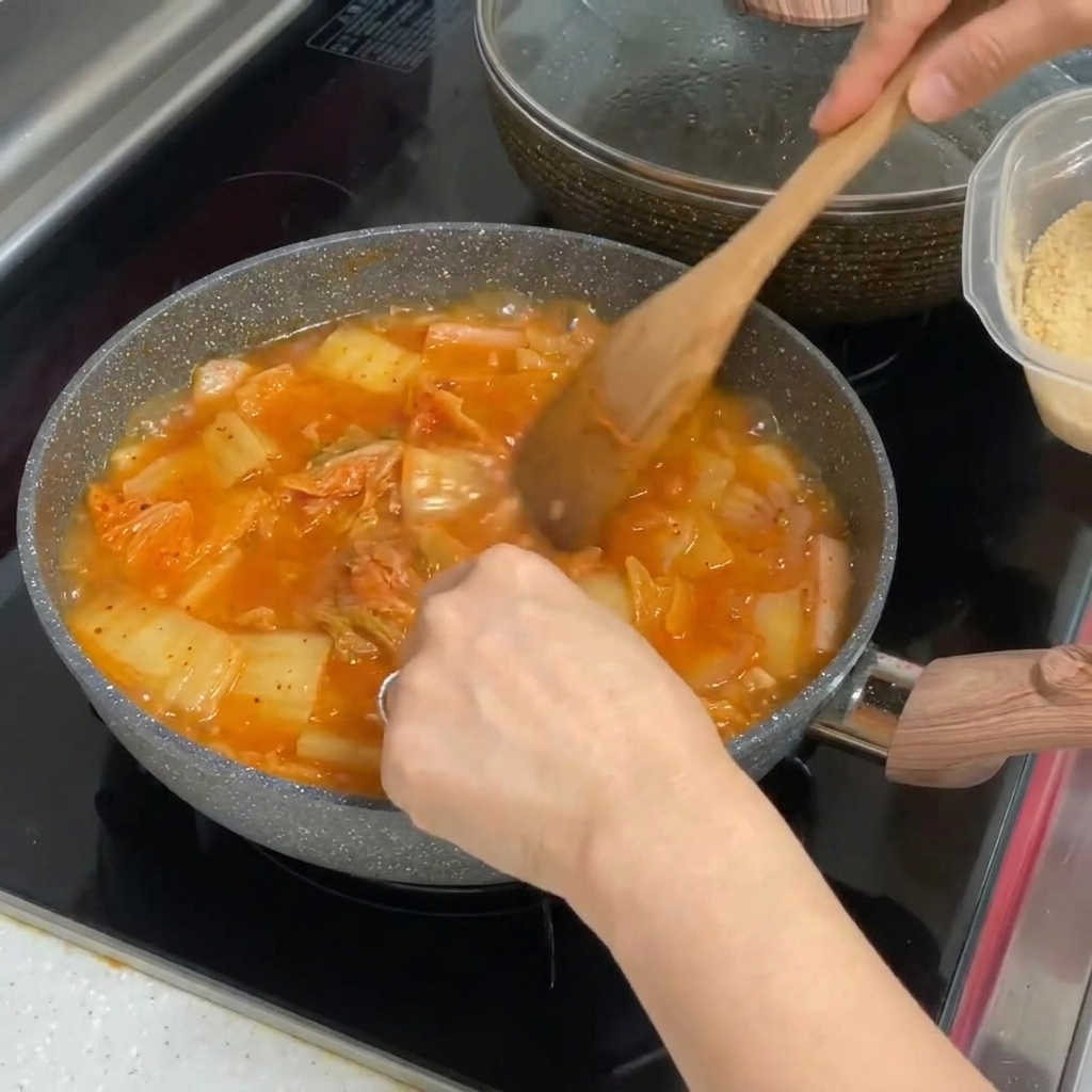 Ajo picado siendo mezclado en una olla de estofado de kimchi burbujeante rojo vibrante.