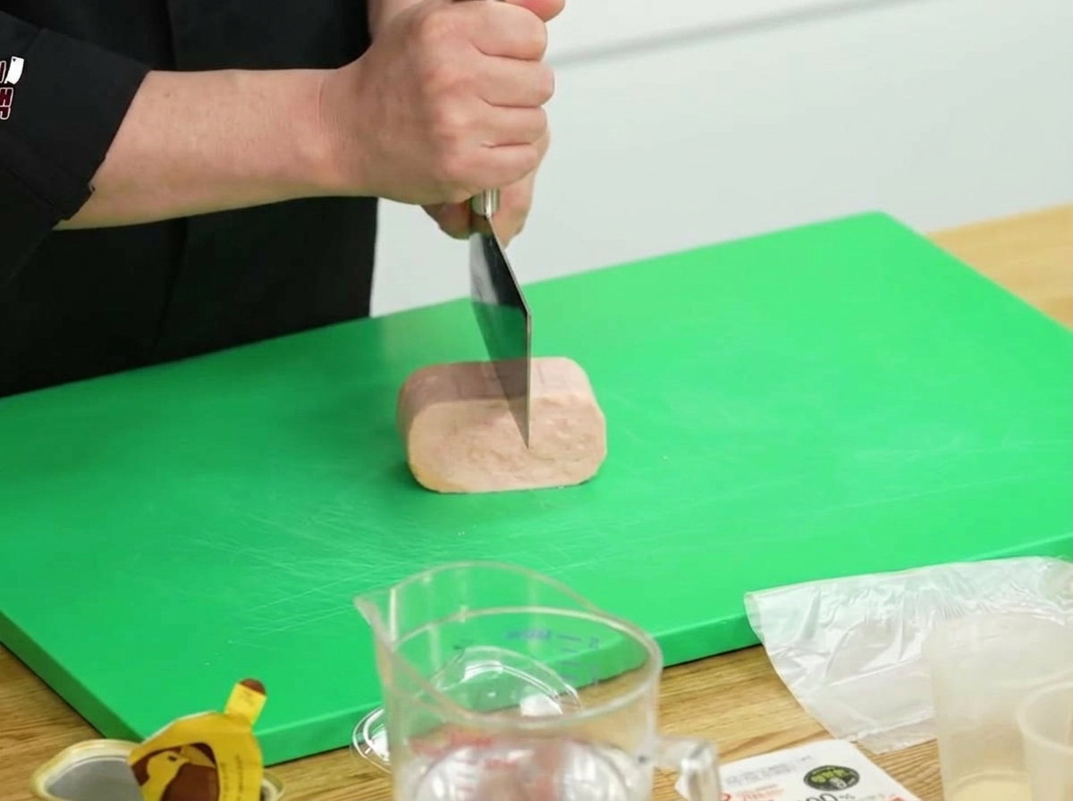 Un bloque de Spam siendo cortado por la mitad con un cuchillo de cocina grande sobre una tabla de cortar verde.