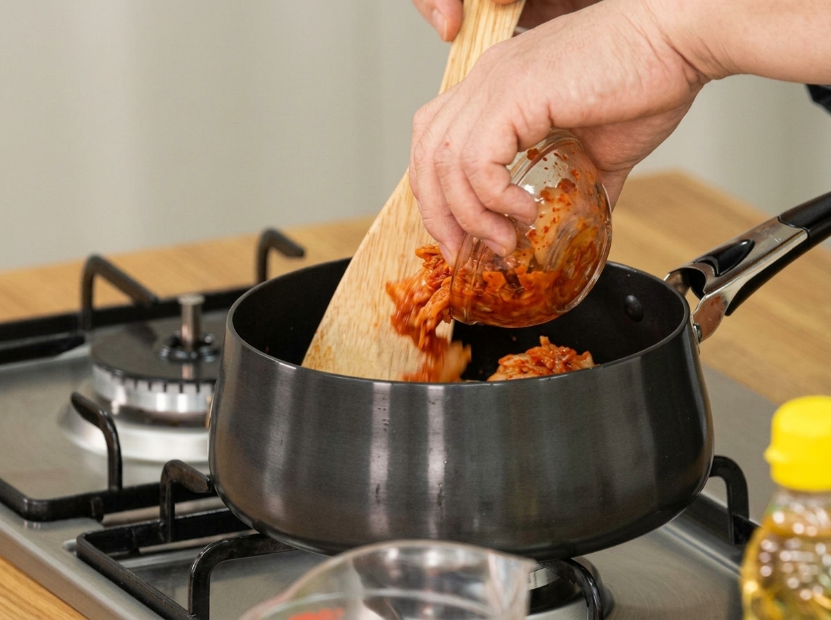 Kimchi añejo siendo añadido a una olla con una cuchara de madera para saltear.
