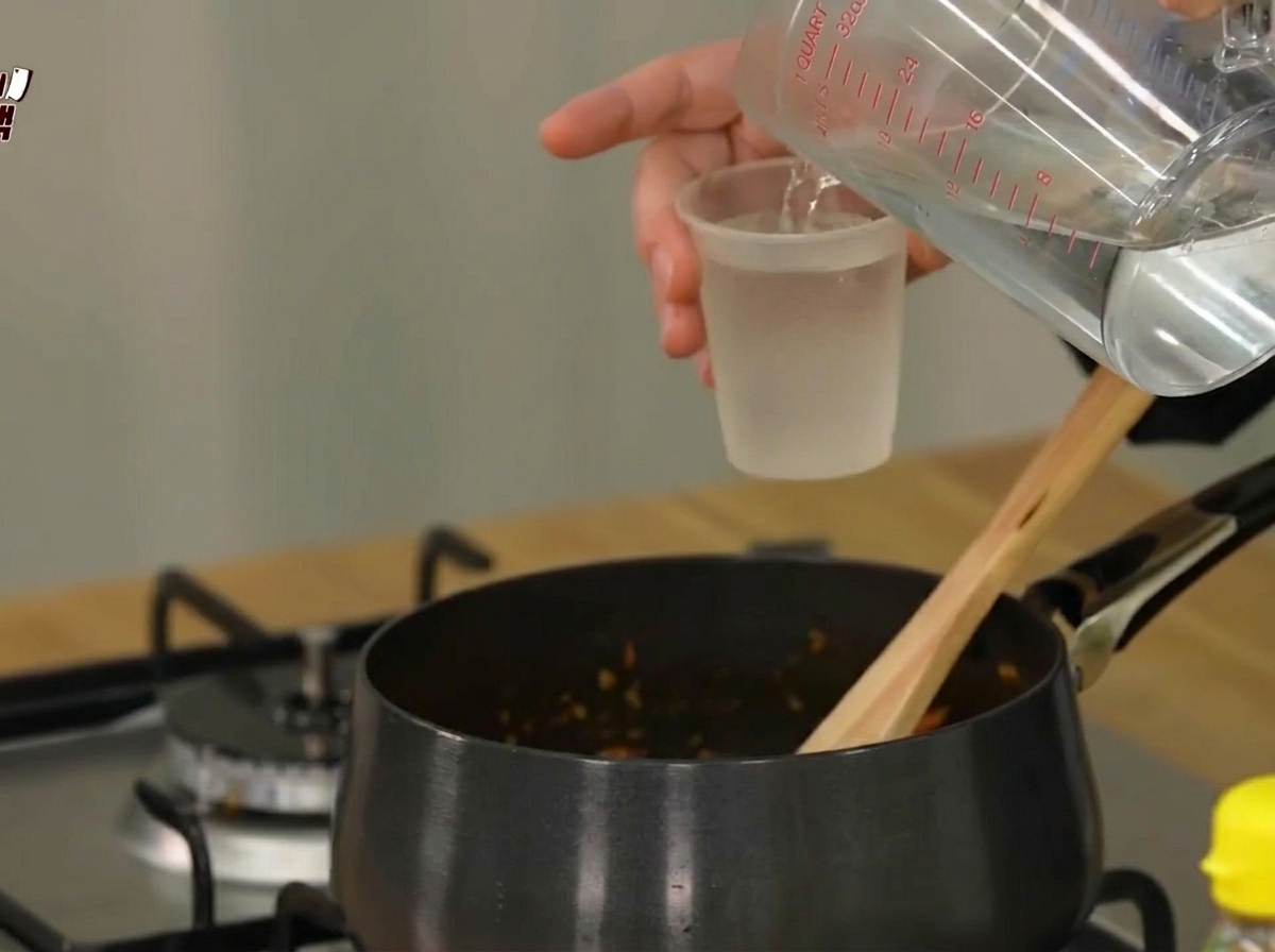 Agua vertiéndose desde una jarra medidora en una olla negra que contiene los ingredientes salteados.