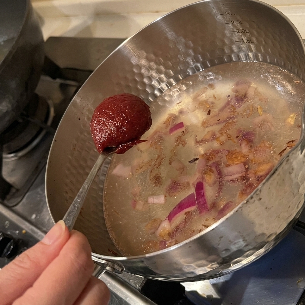 Una cucharada de pasta de chile coreana roja siendo añadida a una olla con agua y cebollas salteadas.