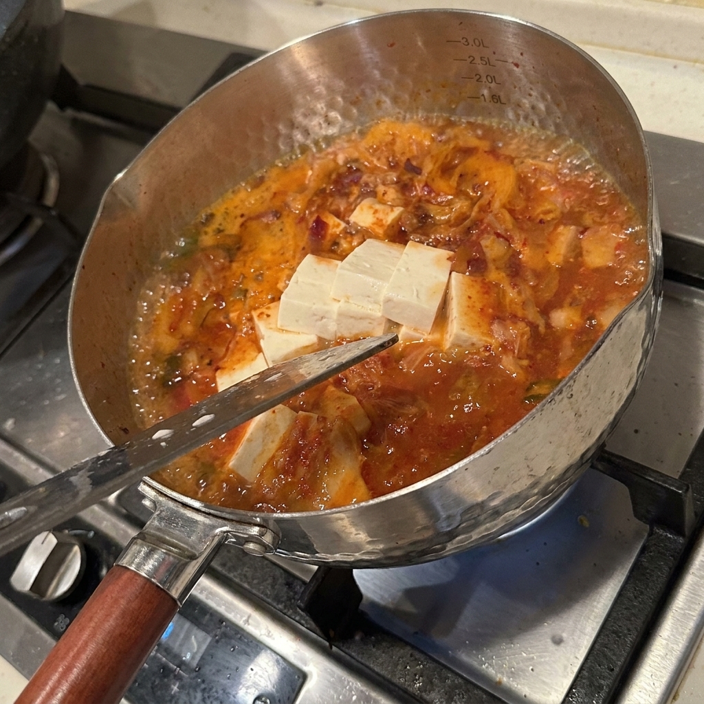 Una variedad de ingredientes frescos incluyendo bloques de tofu, setas enoki y kimchi siendo cocinados a fuego lento en un caldo rojo picante.