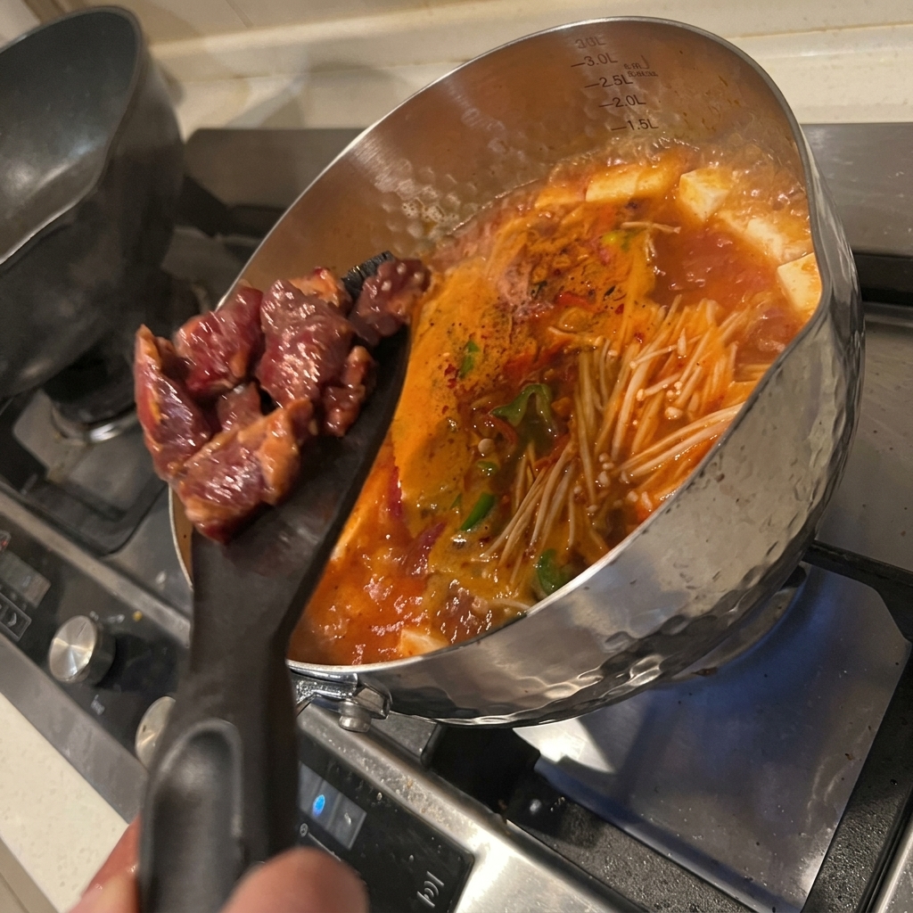 Trozos de ternera marinada siendo añadidos a la olla de sopa de tofu y kimchi hirviendo.