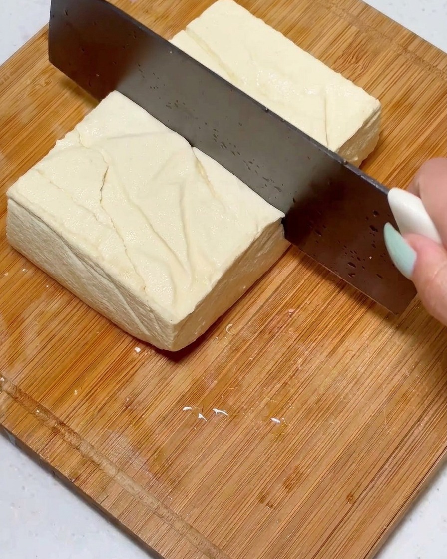 Cortando un bloque cuadrado de tofu firme en trozos uniformes usando un cuchillo grande sobre una superficie de madera.