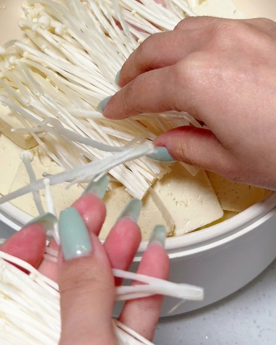 Manos colocando un gran racimo de setas enoki blancas frescas sobre el tofu en rodajas y otros ingredientes en una sartén.