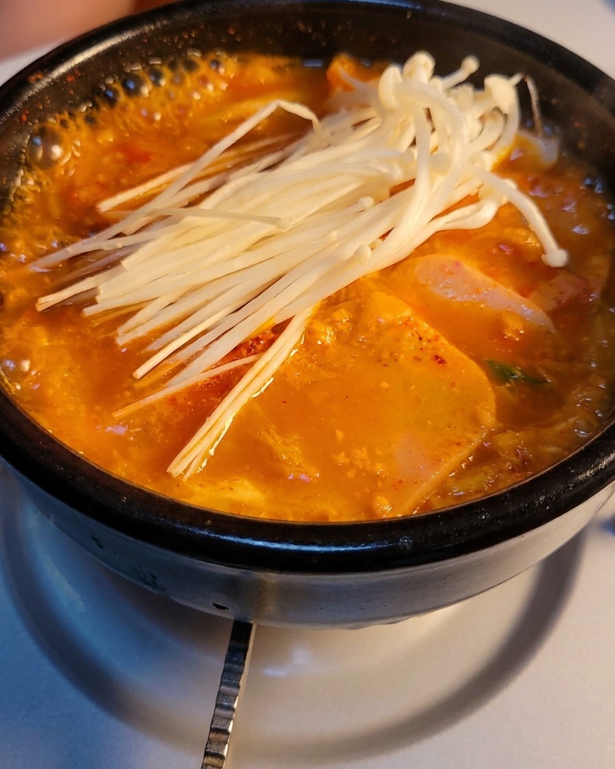 Una olla de piedra con sopa burbujeante de kimchi y carne enlatada, coronada con un puñado de champiñones enoki frescos.
