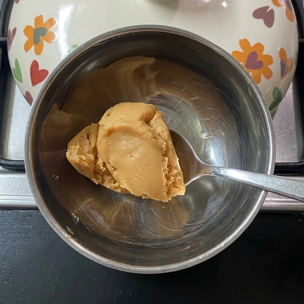 Una cuchara removiendo pasta de miso en agua tibia en un pequeño cuenco de metal.