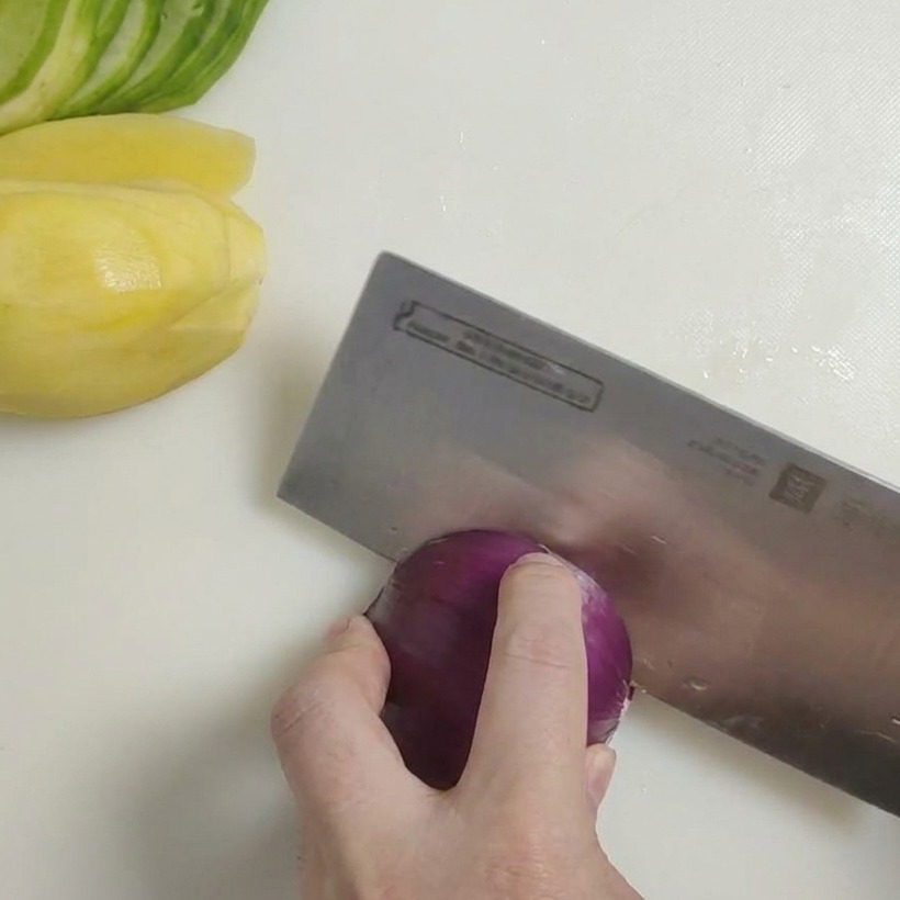 Un cuchillo afilado cortando una cebolla morada en trozos grandes y rústicos sobre una tabla de cortar.