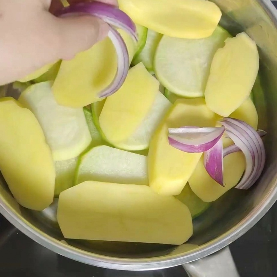 Mano añadiendo trozos de cebolla morada fresca a la olla sobre las patatas.