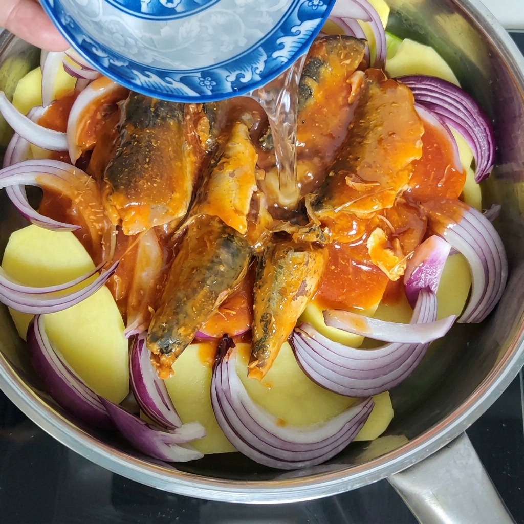 Agua siendo vertida de un pequeño cuenco de cerámica azul y blanco en la olla que contiene la caballa y los vegetales.