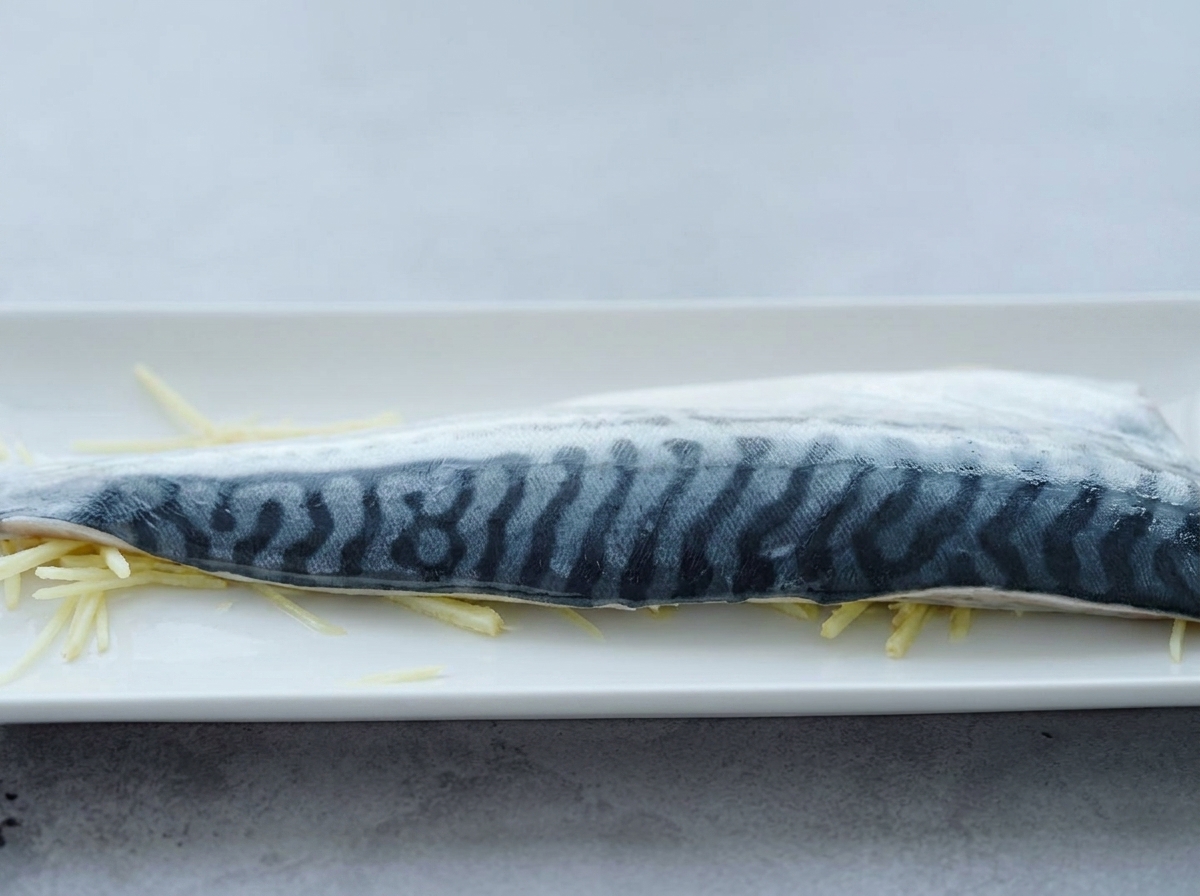 Un filete de caballa cruda sobre una cama de tiras finas de jengibre, con sake de cocina siendo vertido sobre él.