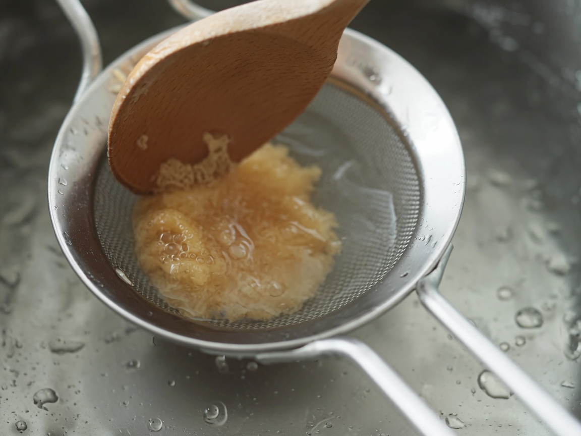 Pasta de miso blanco siendo agitada a través de un colador de metal hacia una sopa hirviendo con una cuchara de madera.