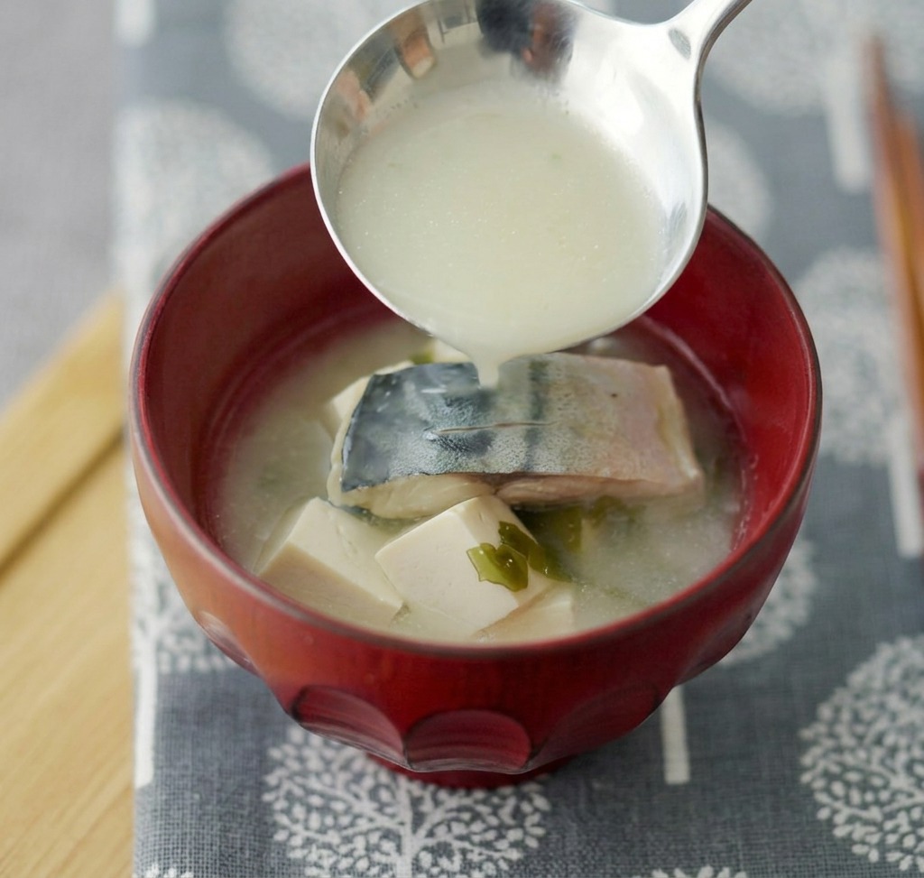 Sopa de miso con caballa y tofu siendo servida con un cucharón en un tazón de cerámica roja profunda.