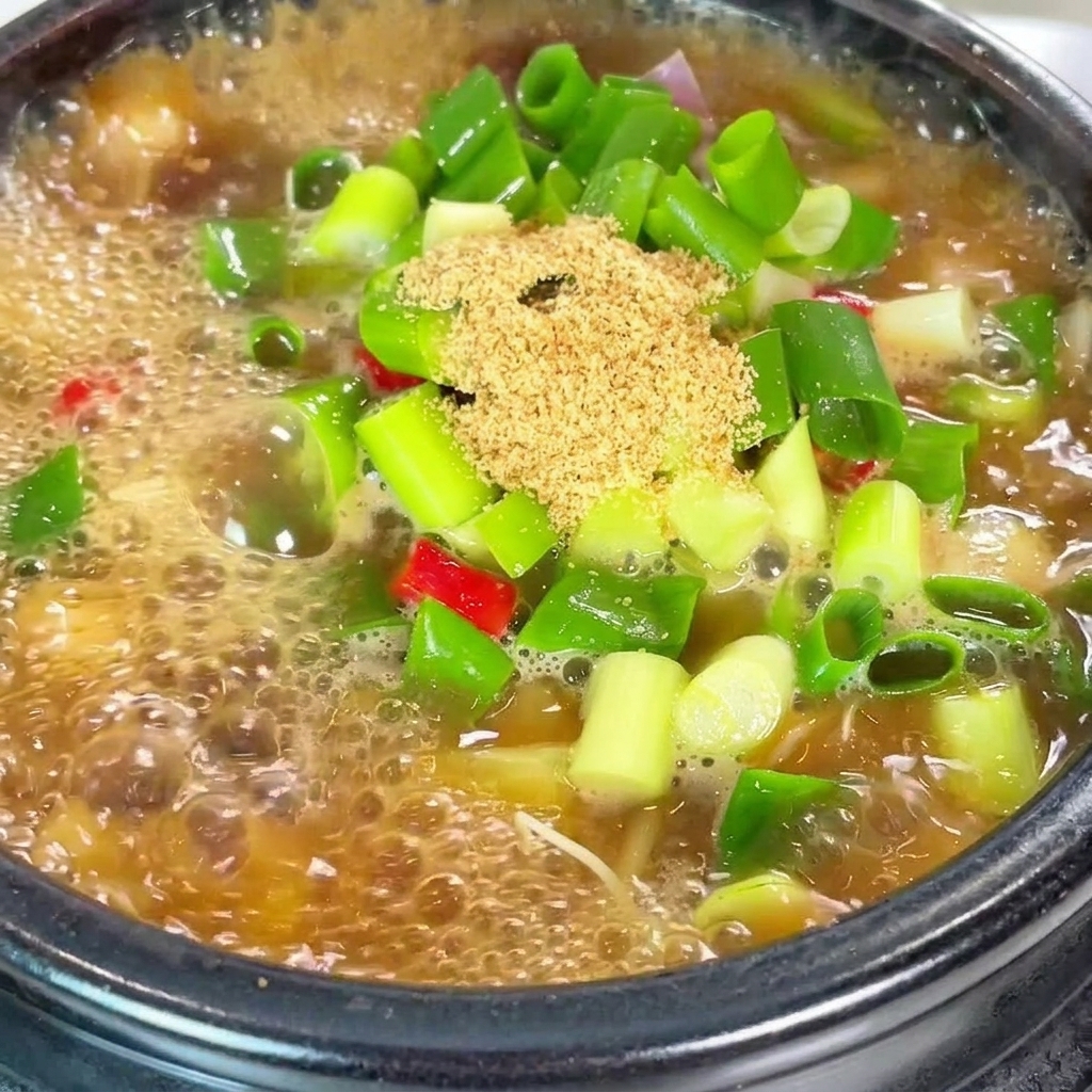 Primer plano del sazonador de carne en polvo y copos de chile rojo brillante siendo añadidos a una olla hirviendo de estofado de soja con cebollino.