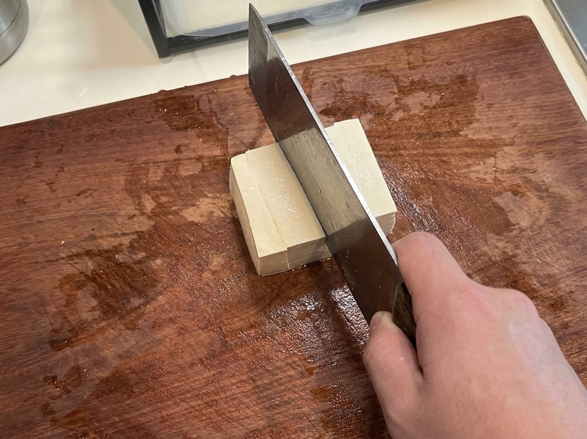 Un bloque de tofu firme siendo cortado en cubos uniformes y limpios sobre una tabla de cortar.