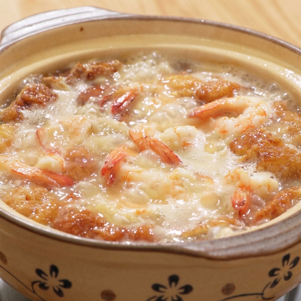 Una olla de barro humeante con sopa de camarones, tofu y huevos, siendo sazonada con pimienta blanca y sal.