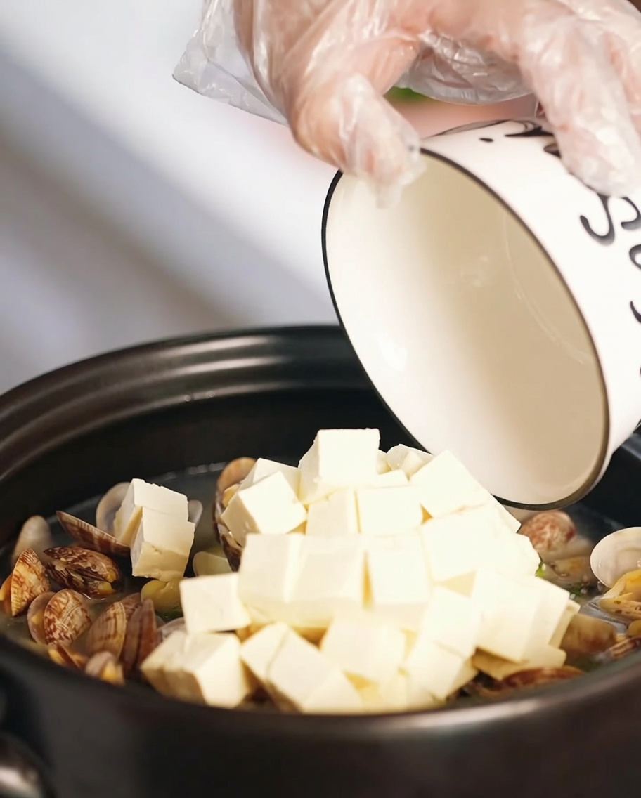 Cubos de tofu suave y trozos de huevo frito siendo añadidos a la sopa de almejas en una olla de barro.