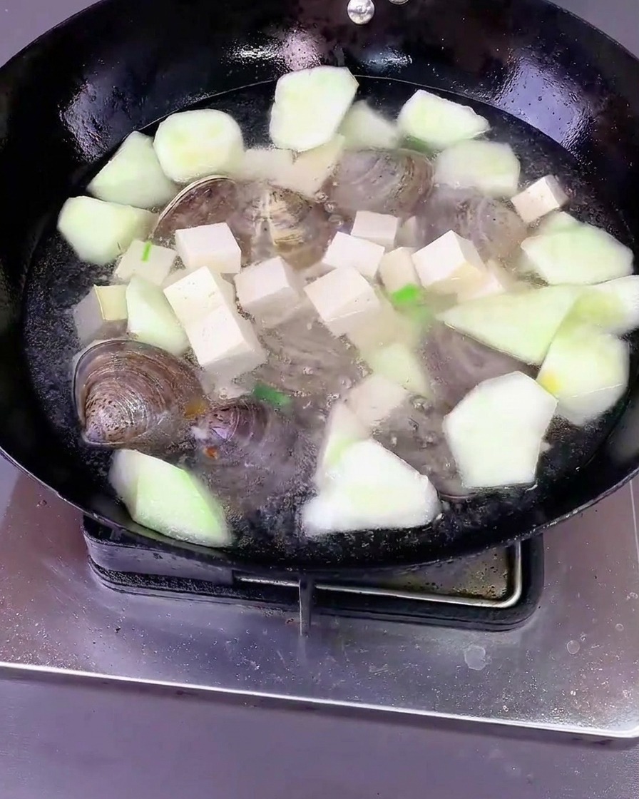Agua vertida sobre la luffa, el tofu y las almejas en un wok, cubierto con una tapa.
