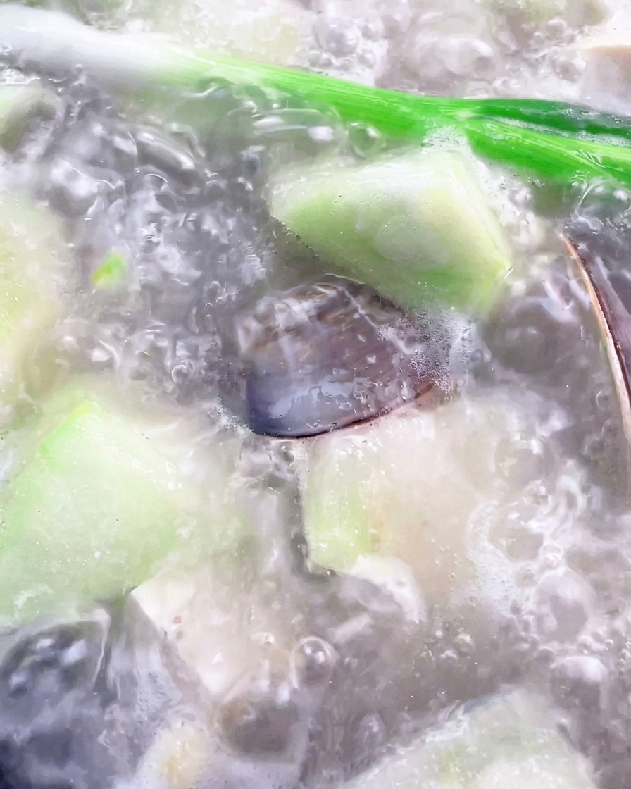 Un cucharón retirando la espuma blanca de la superficie de la sopa de luffa y almejas en una olla.