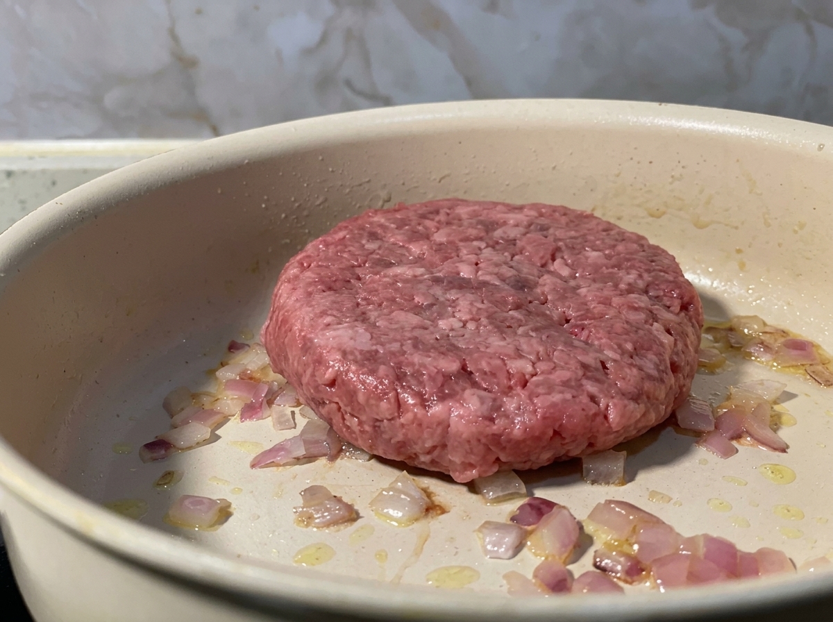 Una hamburguesa de carne picada cruda colocada en una sartén junto a cebolla roja salteada.