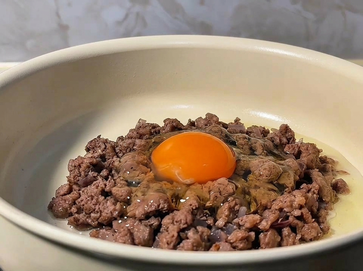 Un huevo crudo cascado en el centro de la carne picada dorada en una sartén.