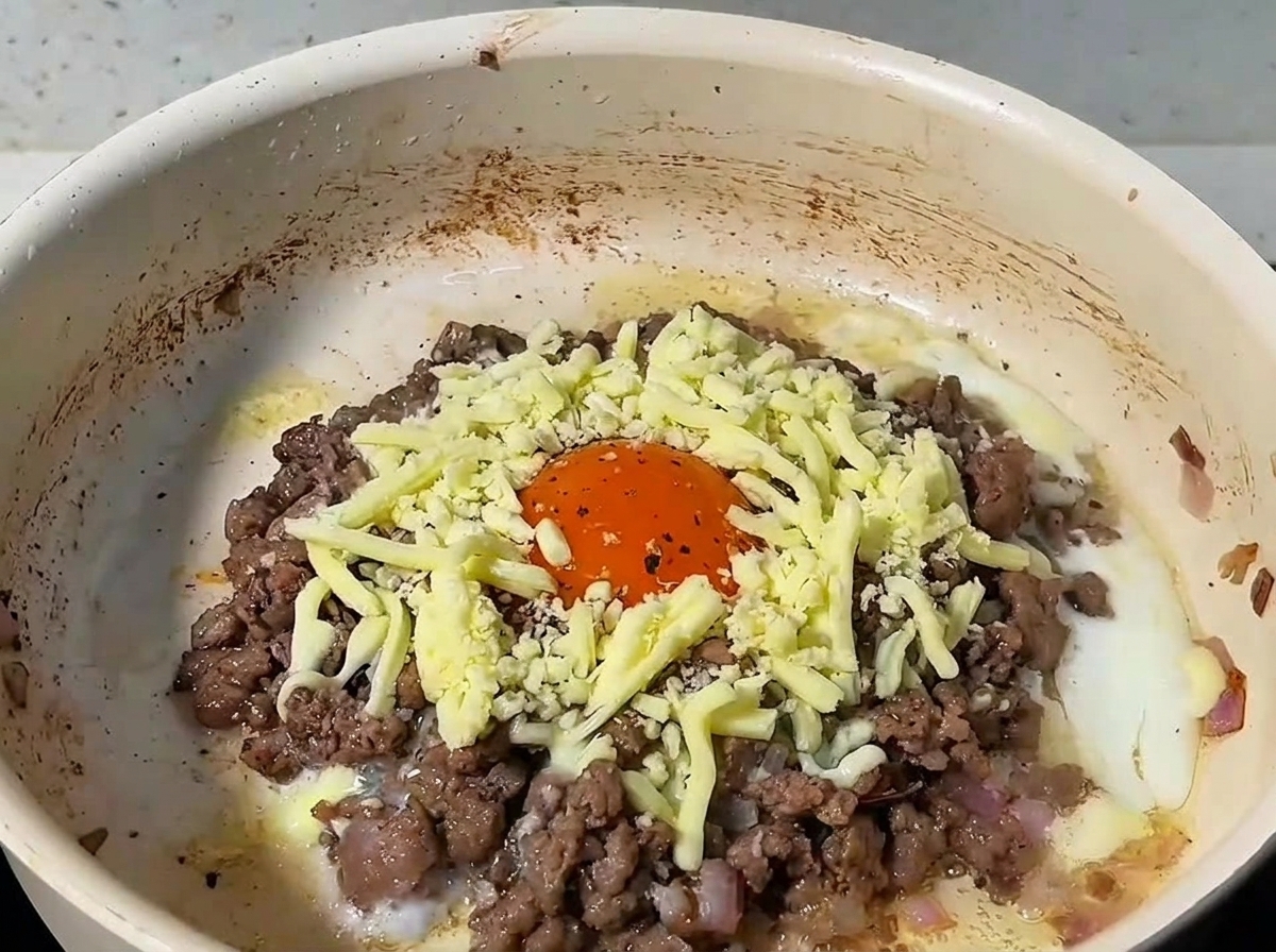 Queso rallado cubriendo la carne picada y el huevo en una sartén, tapada con una tapa.