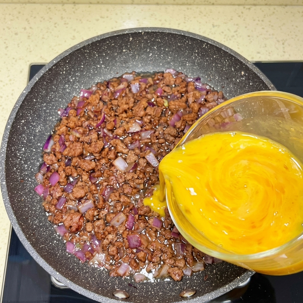 Huevos batidos vertidos desde una jarra medidora sobre una sartén con ternera y cebollas cocinadas.