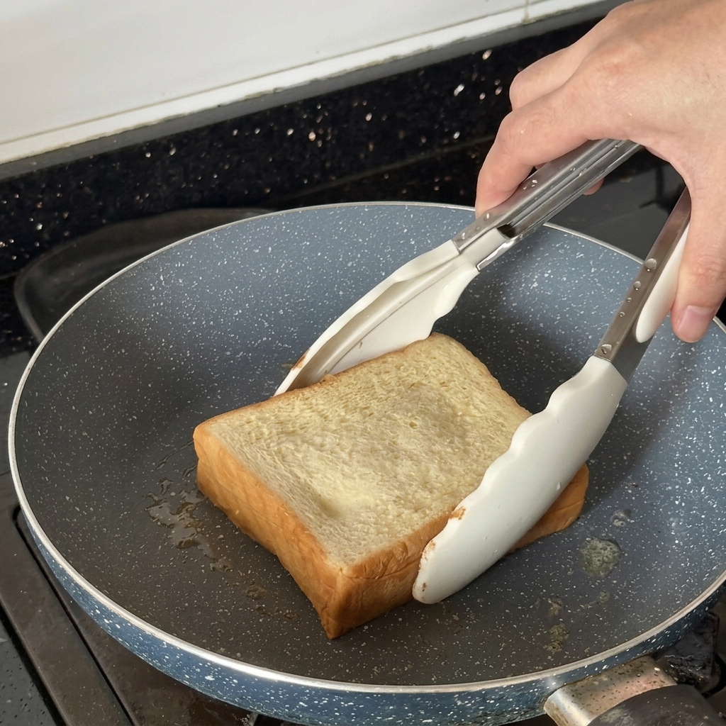 Rebanada gruesa de pan siendo tostada en una sartén usando unas pinzas blancas.