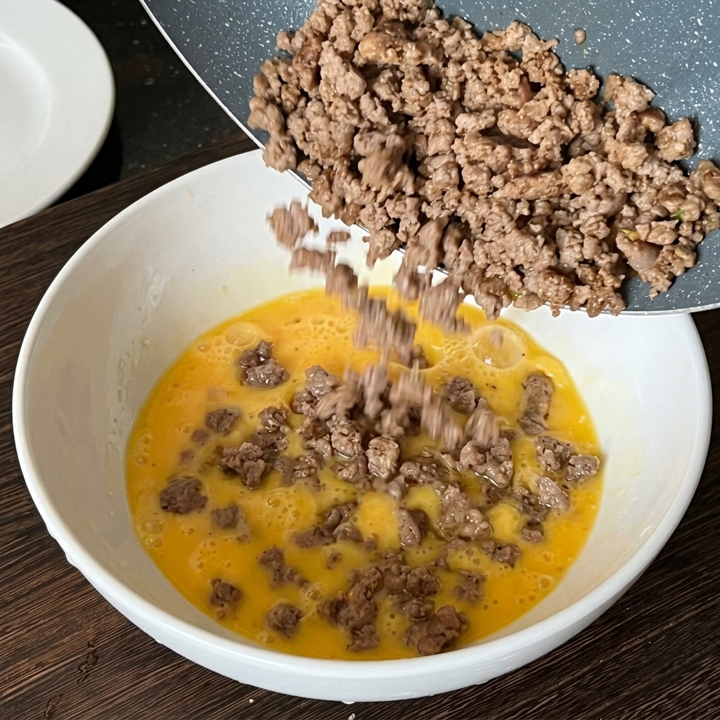 Carne molida cocida siendo vertida desde una sartén hacia un bol con huevos batidos crudos.