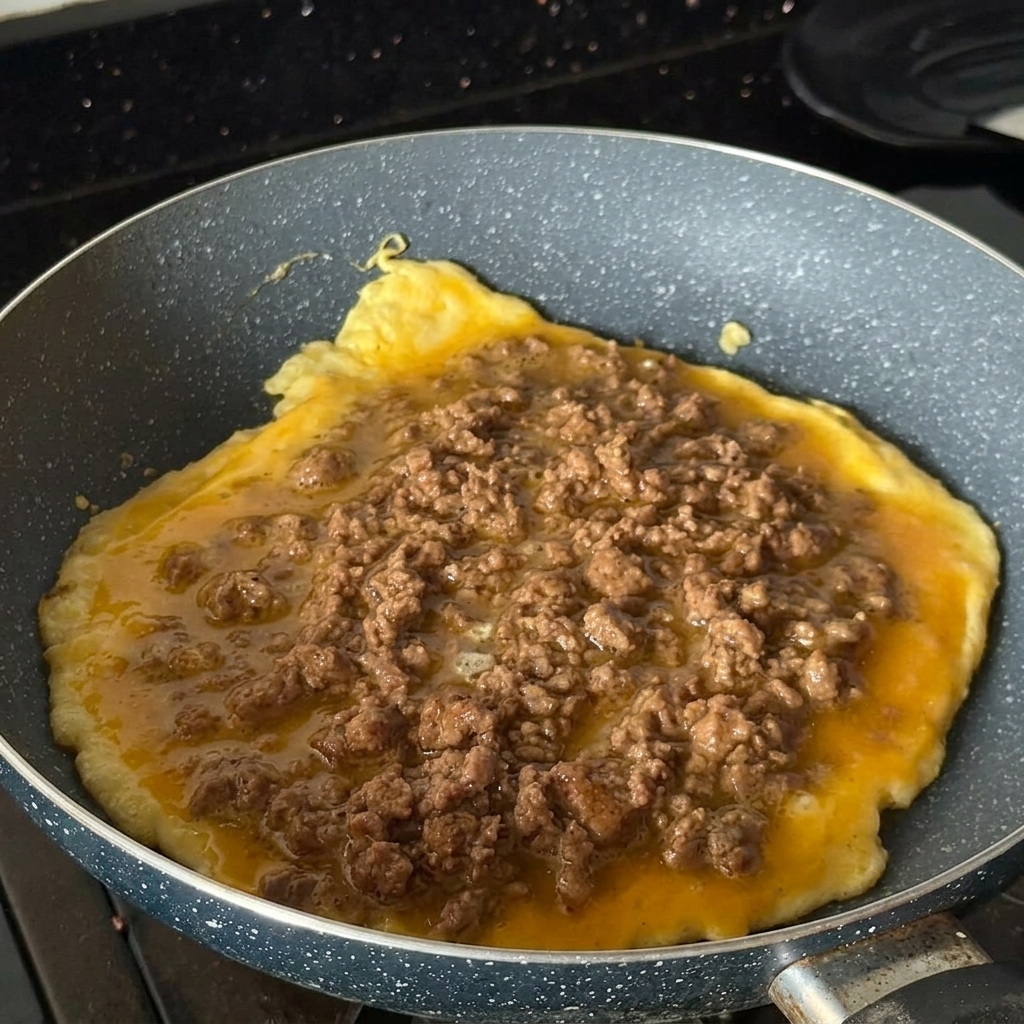 Una tortilla redonda cocinándose en una sartén, cubierta con carne molida sazonada.