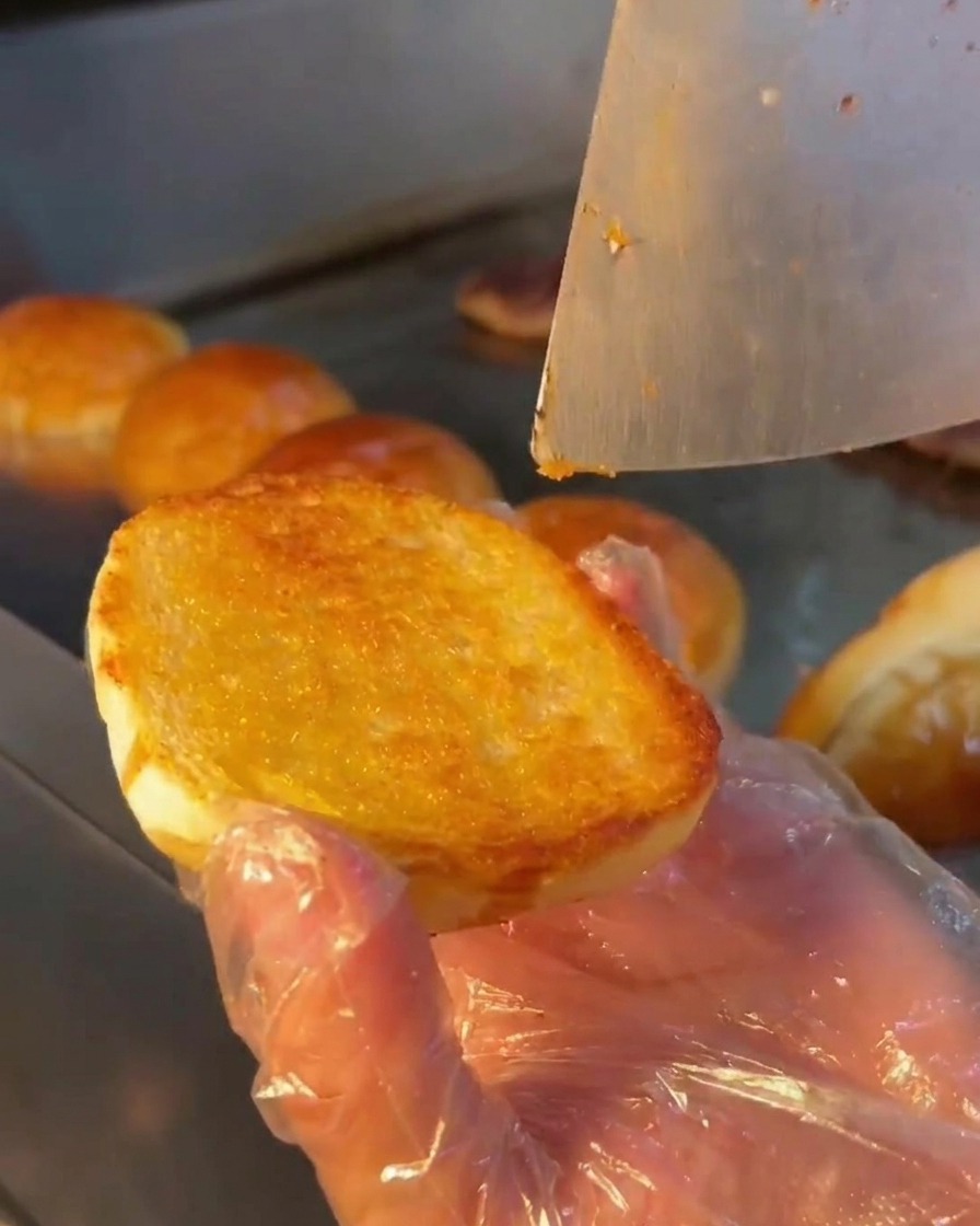Una cuchara de metal extendiendo una salsa blanca cremosa sobre la superficie tostada de un pan de hamburguesa.