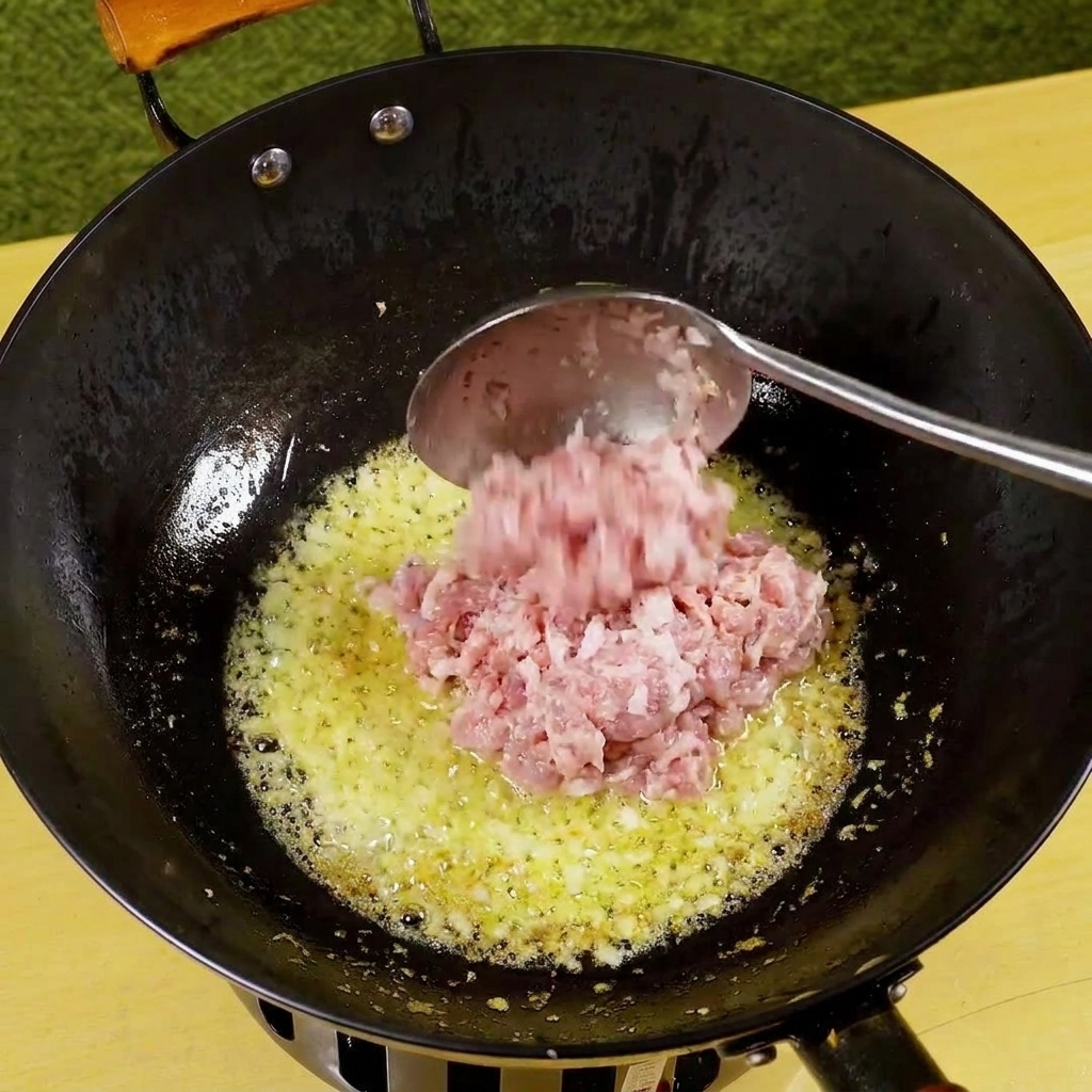 Carne de cerdo picada siendo añadida a una sartén con cebolla y ajo sofritos, comenzando a dorarse.