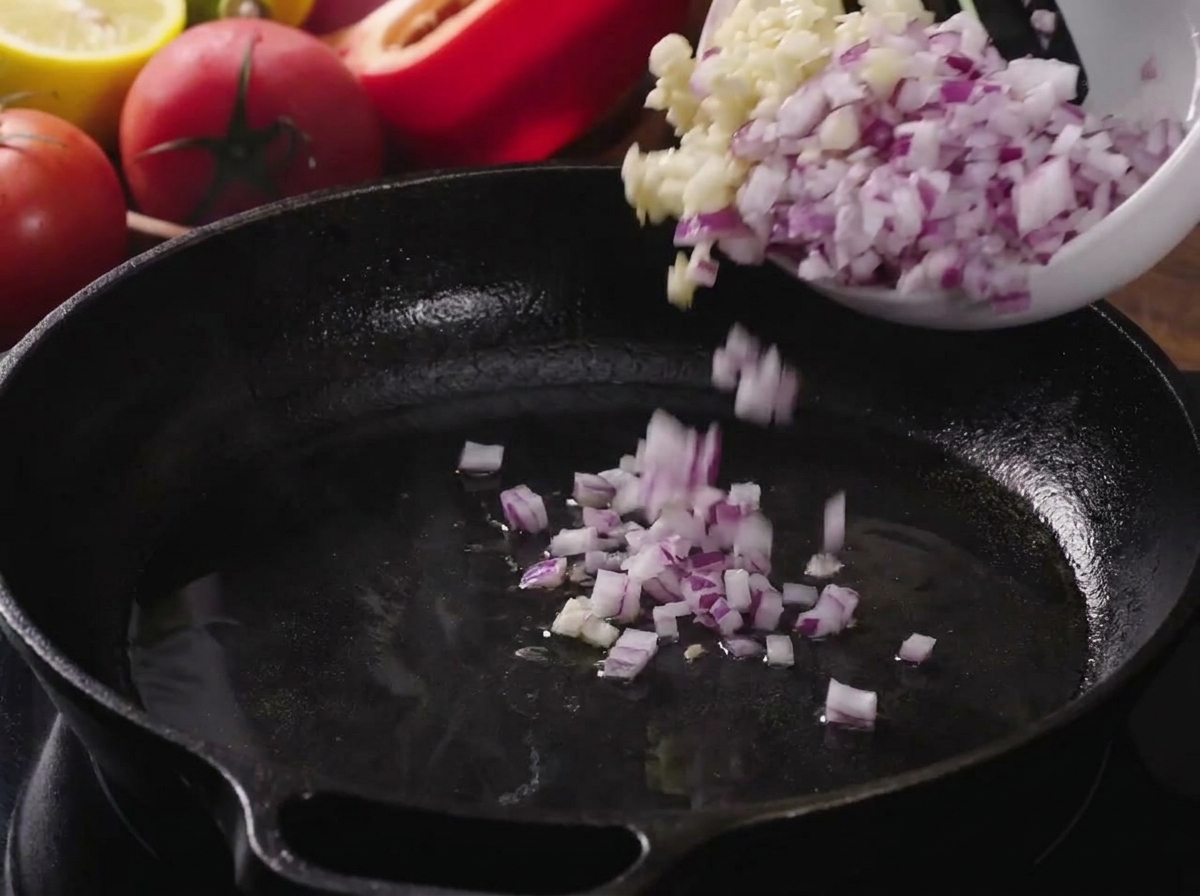 Cebolla picada y ajo picado siendo vertidos en una sartén de hierro fundido caliente con aceite.