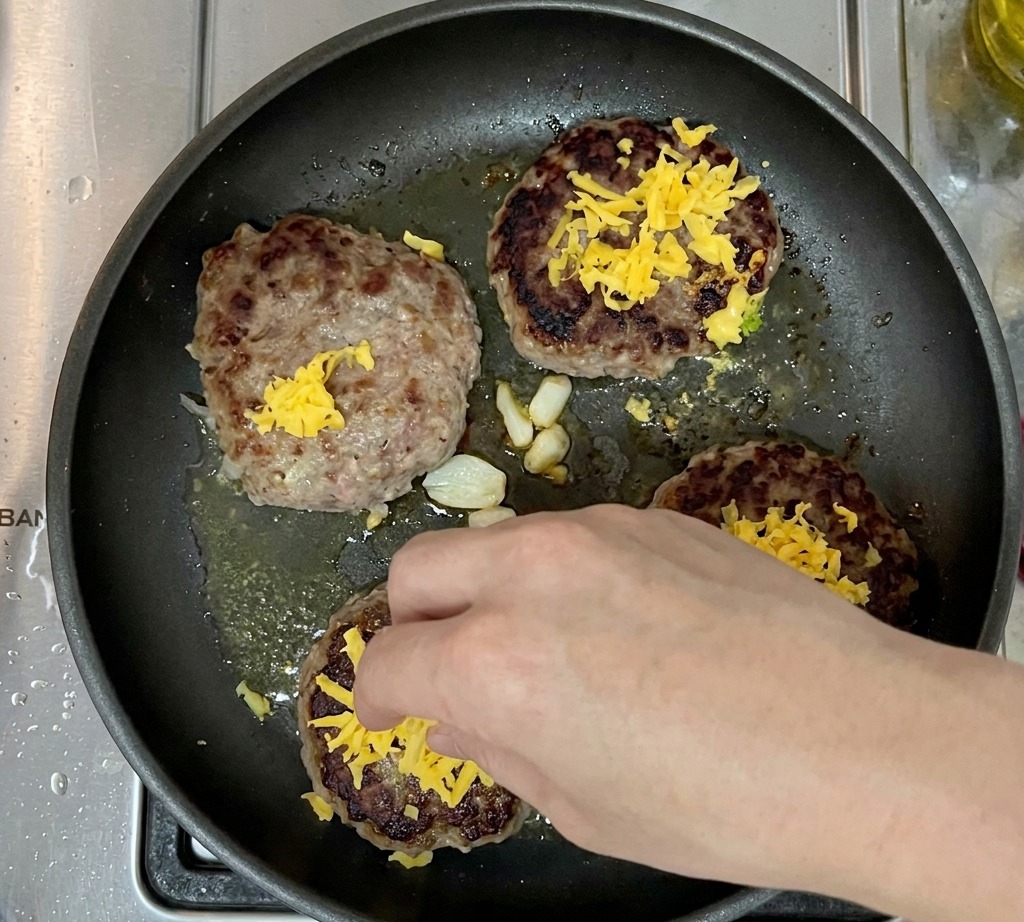 Cuatro hamburguesas cocinadas en una sartén, cada una cubierta con una montaña de queso cheddar rallado.