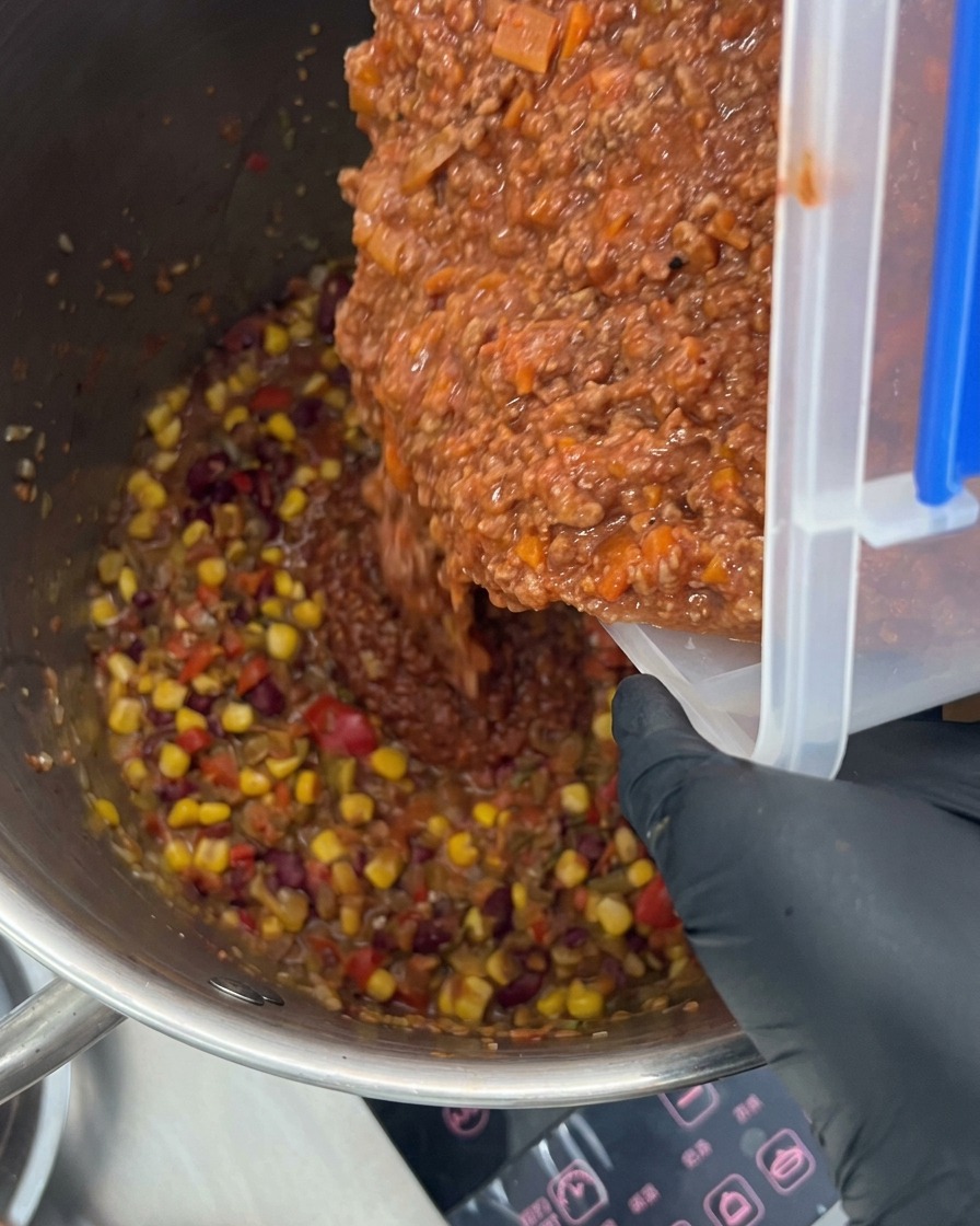 Rica salsa de carne siendo vertida en una olla grande llena de verduras coloridas y especias.