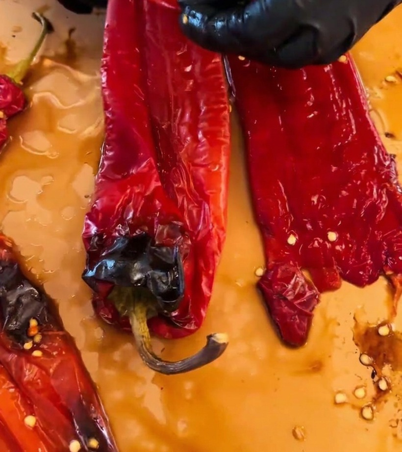 Manos usando guantes negros pelando la piel carbonizada de los chiles rojos asados sobre una superficie de corte.