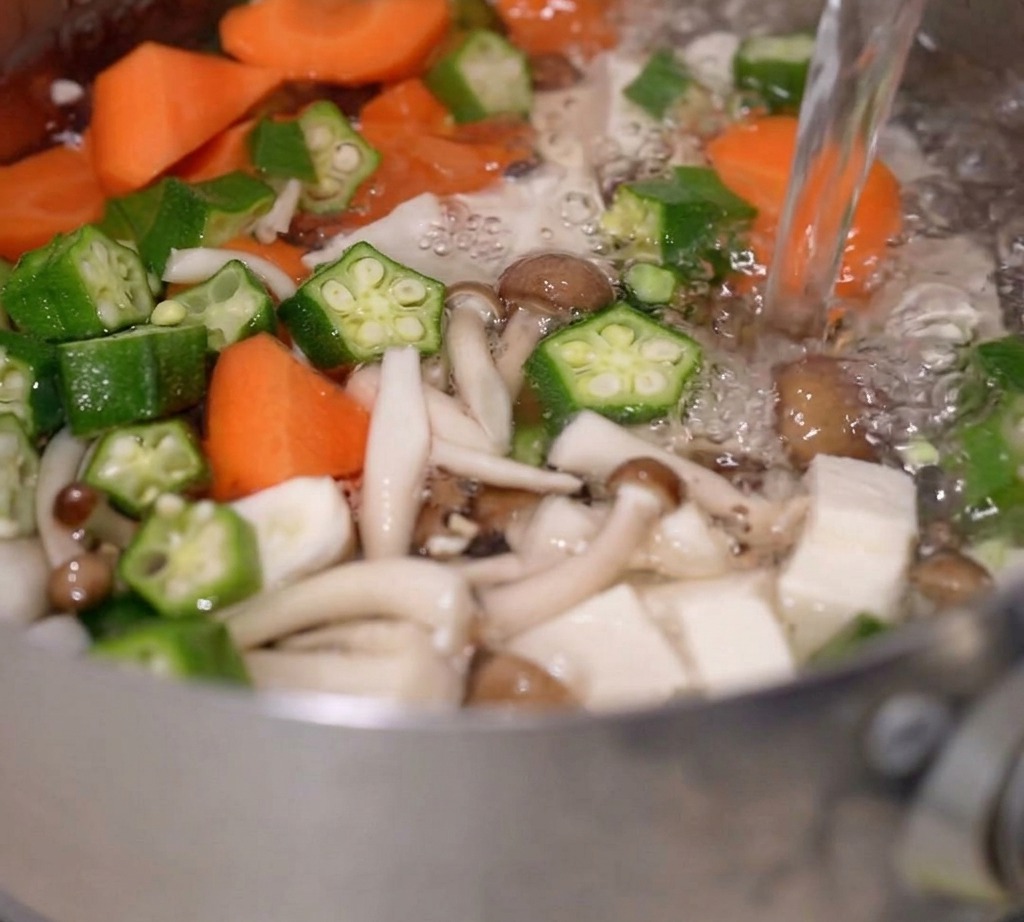 Agua hirviendo siendo vertida en una olla que contiene vegetales mezclados y tofu.