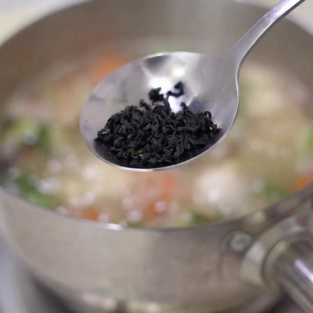 Algas wakame secas siendo añadidas a una olla de sopa hirviendo con zanahorias y otros ingredientes.