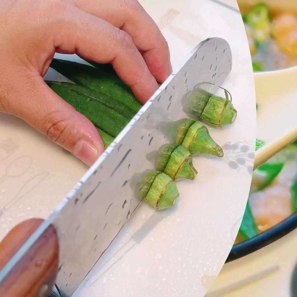 Okra verde fresca siendo cortada cuidadosamente en secciones pequeñas y uniformes sobre una tabla de cortar.