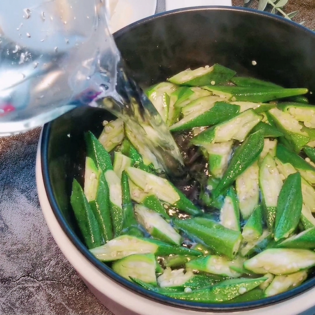 Agua siendo vertida en una olla que contiene trozos de okra salteados.