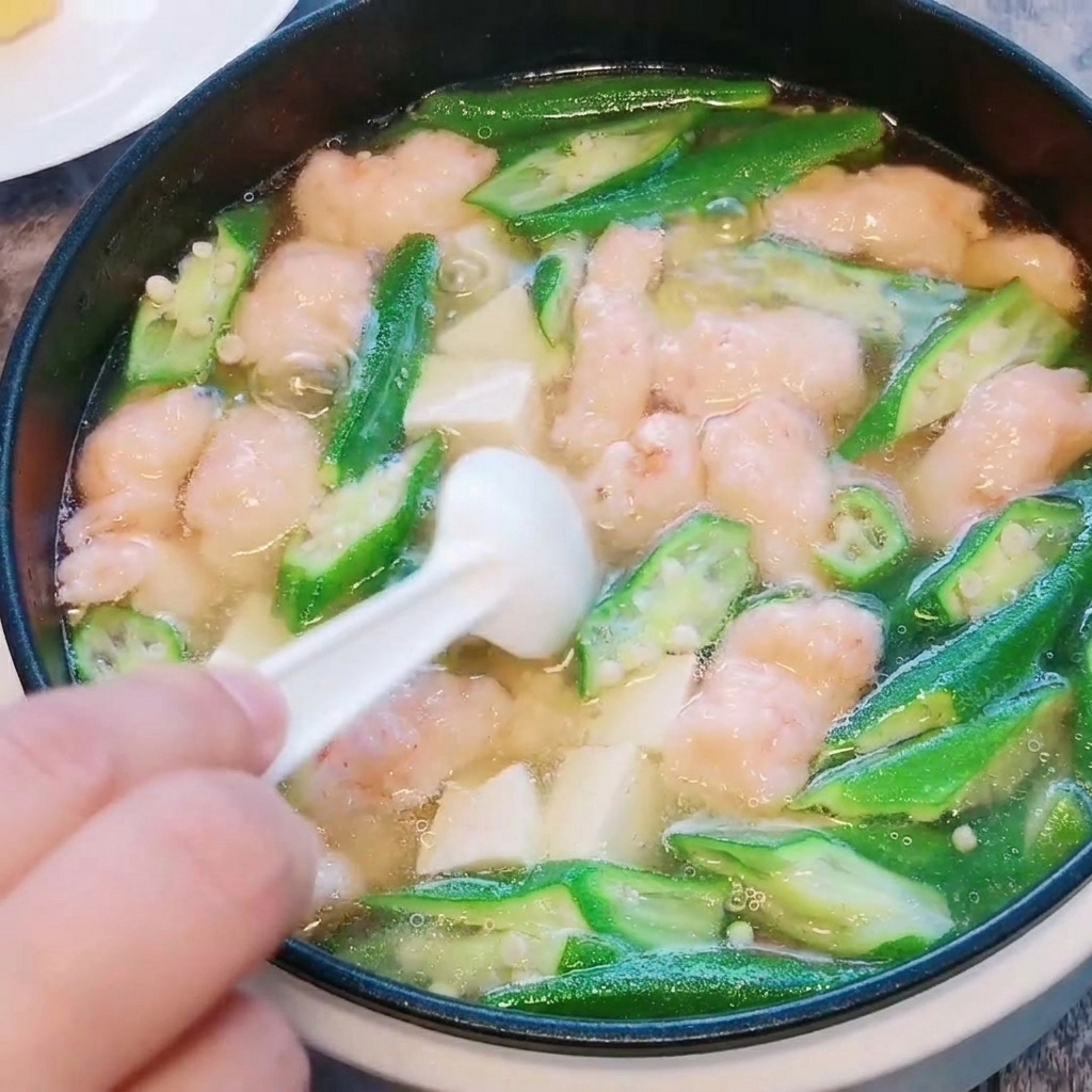Un primer plano de sal y pimienta blanca siendo espolvoreadas en la sopa terminada de okra, tofu y pasta de camarón.